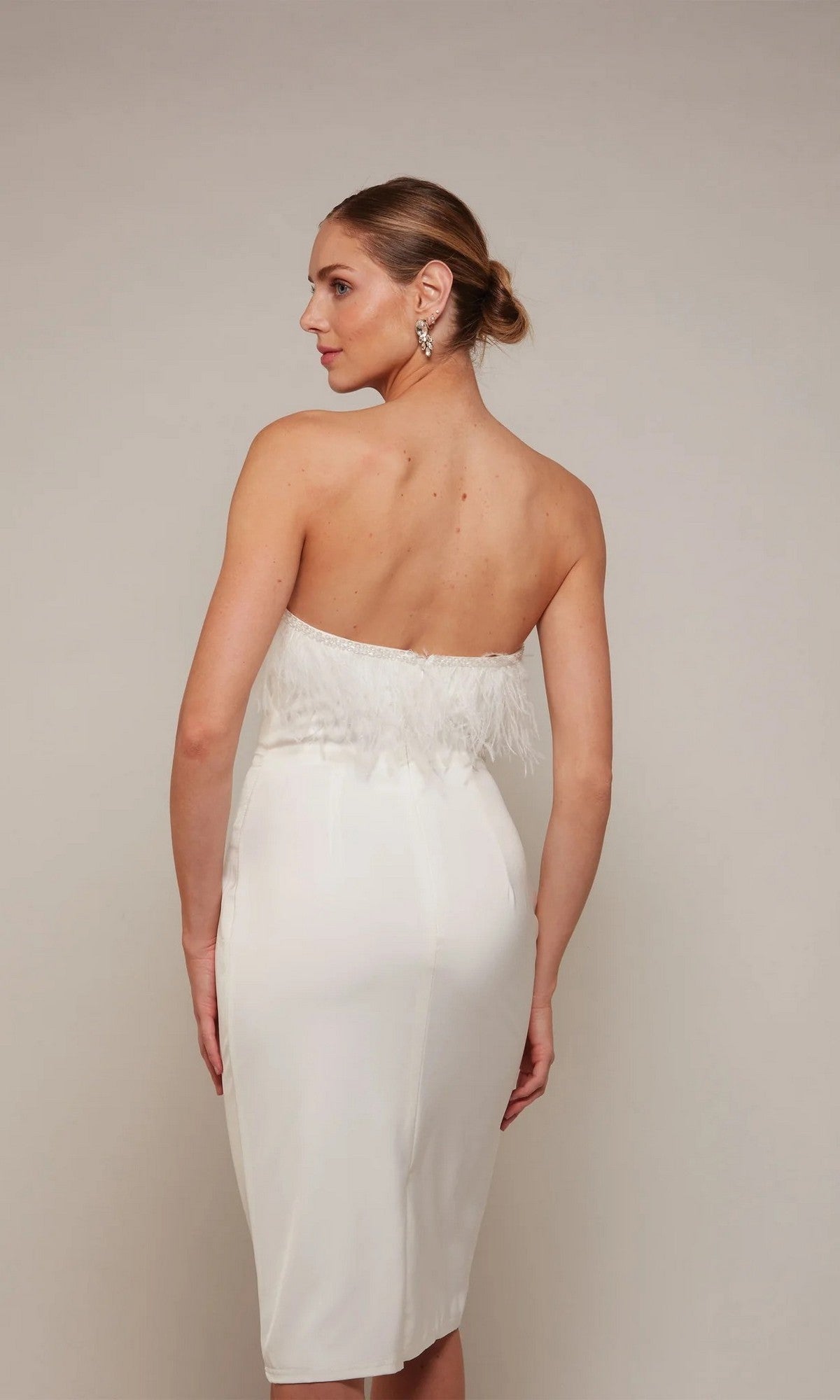 A woman with her hair in a low bun stands facing away against a neutral background, wearing the Alyce 70028 knee-length semi-formal dress featuring a strapless design and feather detailing on the top.
